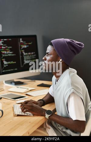 Young serious IT engineer with earphones looking at one of computer screens with coded data while typing on keyboard by workplace Stock Photo