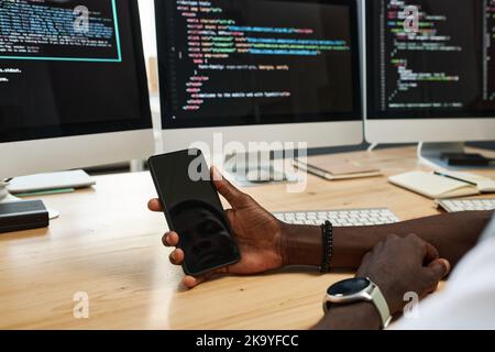 Hands of young African American manager holding smartphone with reflection of his face on blank screen while sitting by workplace Stock Photo