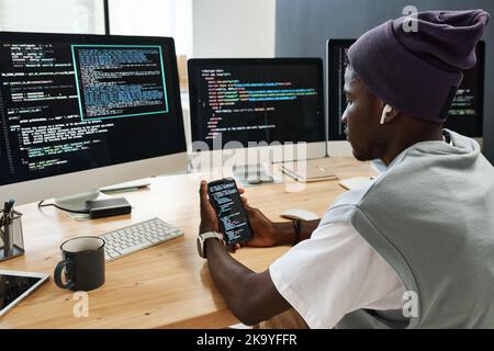 Young serious IT engineer in earphones scrolling through coded data in mobile phone while sitting by workplace in front of monitors Stock Photo