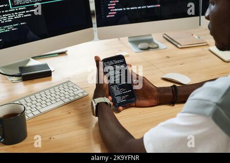 Hands of young black man scrolling through codes in smartphone while sitting by workplace in front of computer screens with data Stock Photo