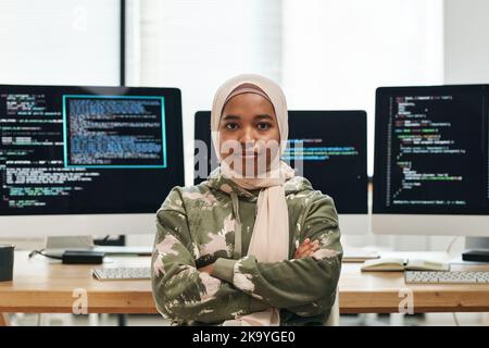 Young successful IT engineer in hijab looking at camera against workplace with computer monitors and coded data on their screens Stock Photo