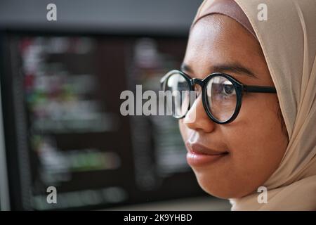 Close-up of face of young confident Muslim intern in hijab and eyeglasses standing in front of camera against computer screen with data Stock Photo