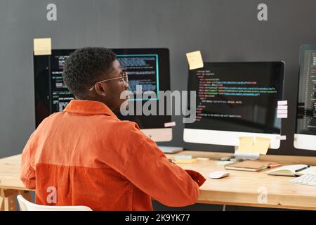 Rear view of young black man concentrating on decoding data on computer screen while sitting by workplace in front of monitors Stock Photo