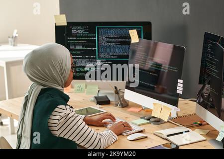 Young woman in hijab typing on computer keyboard while sitting by workplace in front of monitor in office and decoding data Stock Photo