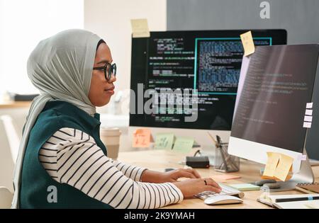 Young Muslim businesswoman in hijab looking at computer screen while typing and decoding data in front of monitor in office Stock Photo