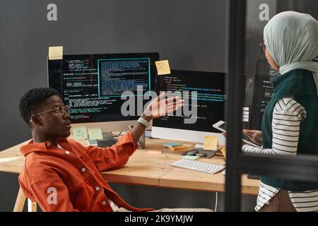 Young confident African American man making presentation of decoded data to female colleague in hijab listening to him at meeting Stock Photo