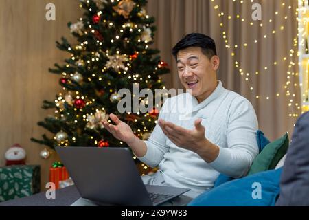 Happy christmas man making video call using laptop sitting on sofa in living room at home, asian man greeting friends online happy new year smiling and talking happily. Stock Photo
