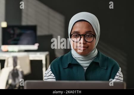 Young IT support engineer in casualwear, eyeglasses and hijab sitting in front of computer monitor and networking in office Stock Photo