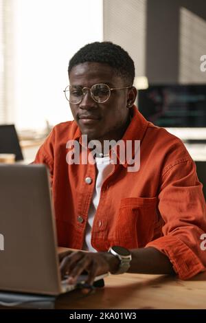 Serious young IT support engineer in casualwear and eyeglasses sitting by workplace in front of laptop and networking or decoding data Stock Photo