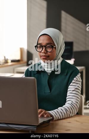 Young intercultural businesswoman in hijab and eyeglasses sitting in front of laptop and decoding data or creating new website Stock Photo