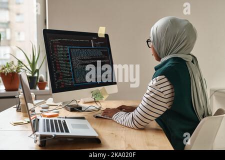 Young Muslim businesswoman or intern looking at computer screen while typing and decoding data or developing software by workplace Stock Photo