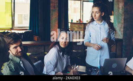 Confident young woman executive director is talking to multi-ethnic group of co-workers asking questions and giving orders during meeting in modern office. People and business concept. Stock Photo