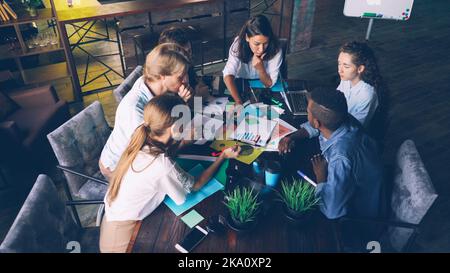 High angle view of multiracial team working with graphs and charts sitting at table, looking at papers and discussing business strategy. Laptop, to-go coffee and documents are visible. Stock Photo