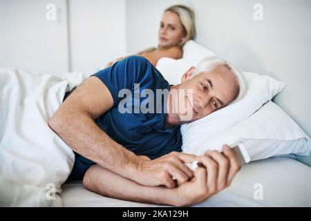 His happiness just turned into her concern. a mature man using a cellphone with his wife looking upset in the background at home. Stock Photo