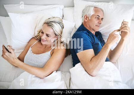 Caught up with their own connections. a mature couple using their cellphones in bed at home. Stock Photo