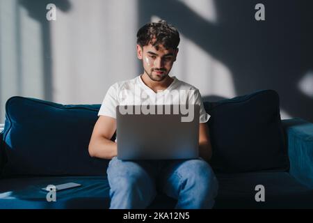 Attractive young man relaxing on a couch at home, holding, using and working on laptop pc computer spending his free spare time in living room Stock Photo