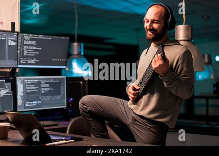 App developer singing and playing guitar with keyboard, using IT equipment and fooling around. Silly funky programmer celebrating success, wokring on server code after hours in office. Stock Photo