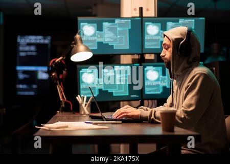 Male criminal trying to break database server, using virus to code malware and steal big data at night. Working with multiple monitors and hacking information system, wearing hood. Stock Photo
