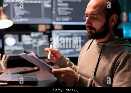 Software developer taking notes on files after comparing html script and source code, doing online database analysis. Writing coding information on papers, working late at night in office. Stock Photo