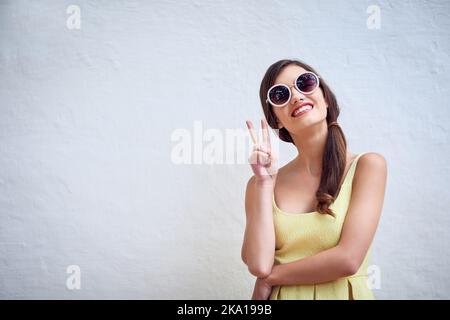 She comes in peace. Studio portrait of a cheerful young woman wearing sunglasses while showing a peace sign against a grey background. Stock Photo
