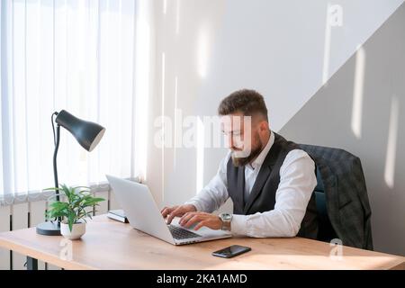 Caucasian man sits at table near laptop in office and looks at screen. Serious and thoughtful bearded boss or employee working at his workplace and feeling interested or analyzing the results. Stock Photo