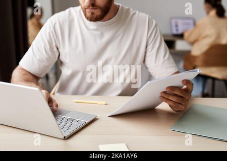 Close-up of young man in white t-shirt holding copybook and using laptop while sitting by desk during online lesson with tutor Stock Photo