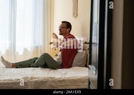 Man in wireless earbuds sitting on bed with laptop listening favorite music online and gesturing Stock Photo