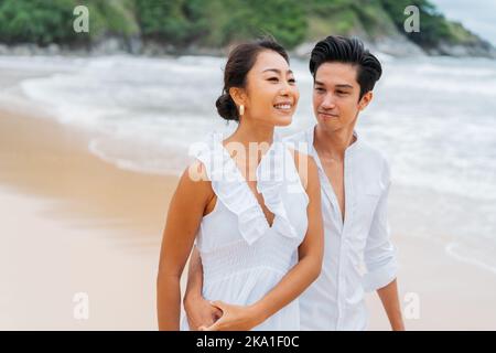 Happy romantic Asian 30s couple in relationship holding hands on the sandy beach in summer. A smiling couple wearing a white shirt and dress in daytime. Stock Photo