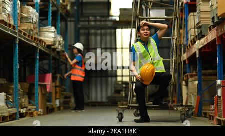 Tired male warehouse worker sitting and wipe the sweat away, taking break from hard work in large warehouse Stock Photo