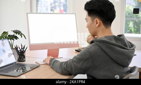 Smart and hipster young Asian male programmer or office worker working at his desk, using PC computer and notebook laptop, planning his project. compu Stock Photo