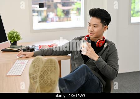 Hipster and chill young Asian male programmer or web developer enjoys his coffee and puts his legs on the table while working in the office. Stock Photo
