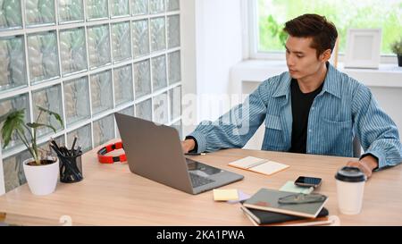 Handsome and smart millennial Asian male programmer or freelancer in jeans jacket, sits at his desk, works on his project on laptop computer. Stock Photo