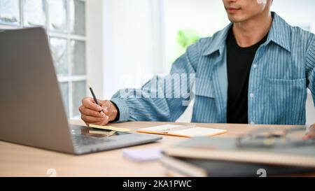 cropped image, Smart and good looking young Asian college student or male freelancer in jeans jacket working on his project, taking notes in his noteb Stock Photo