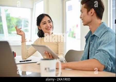 Happy and charming young Asian female having a good conversation with her male colleague while working together in the office. Stock Photo