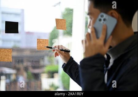 close-up image, Young Asian male office employee is on the phone with his colleague while noting something on the memo sticky note on the glass wall. Stock Photo