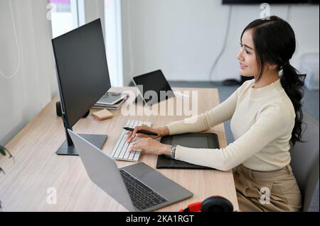 Charming and attractive young Asian female web developer or graphic designer working at her desk. computer, laptop and stylus tablet are on the table Stock Photo