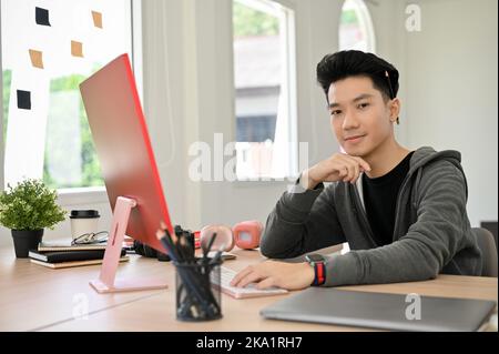 Portrait, Handsome and smart young Asian male programmer to web developer in casual clothes sitting at his modern office desk, smiling, hand on chin, Stock Photo