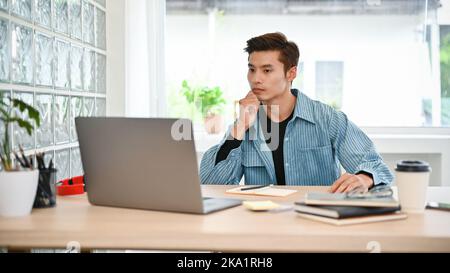 Handsome young Asian male college student or office employee focused on thinking and planning his project, using laptop computer, remote working at th Stock Photo