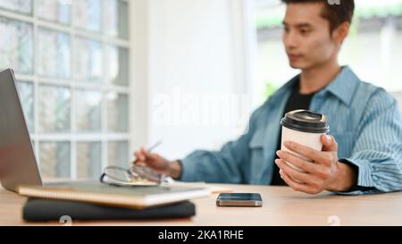 selective focus hand image, Handsome millennial Asian businessman in casual clothes holding a coffee cup while working on laptop. Stock Photo