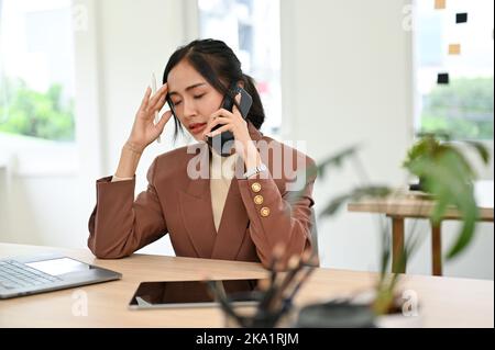 Stressed millennial Asian businesswoman or female manager sitting at her desk, having a serious phone call with her business partner. Stock Photo