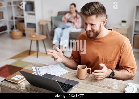 Young smiling man in casualwear sitting in front of laptop at home and communicating with his tutor during online lesson Stock Photo