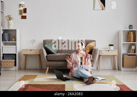 Young smiling woman in headphones sitting by couch on the floor in front of laptop and communicating with tutor during online lesson Stock Photo