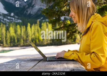Smiling woman using her laptop with amazing view of mountains. Freelancer wearing yellow jacket and working remotely in the forest.  Stock Photo
