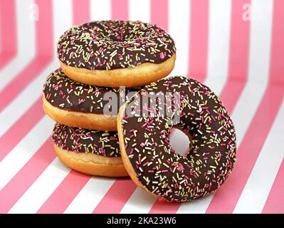 stacked doughnuts with chocolate icing and colored sprinkles on white pink striped background, close up of delicious donuts Stock Photo