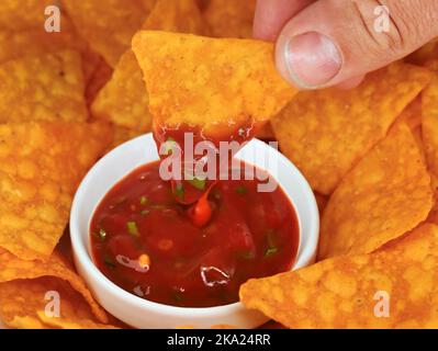close up of dipping a tortilla chip in hot spicy salsa with chili peppers, classic mexican corn chips as snack for partys served in a bowl Stock Photo