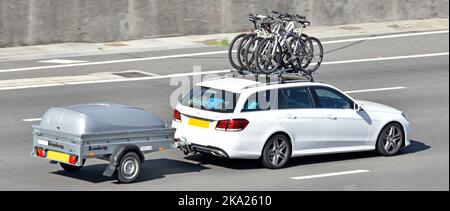 Roof rack fixed on white Mercedes car side view transporting cyclists bikes and towing a covered Brenderup 1205 trailer driving on UK motorway road Stock Photo