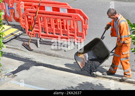 Road works workman wearing high visibility safety clothes tipping backfill tarmac out of wheelbarrow reinstating pavement broadband cable trench UK Stock Photo
