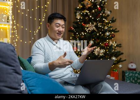 Quarantine for holidays. Young handsome Asian man sitting at home on sofa alone on New Year near Christmas tree. Holds a laptop, talks to family, friends via online video call. Stock Photo