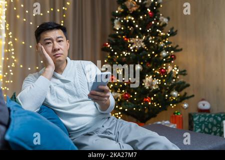 Sad and lonely young Asian man sits at home on the sofa near the Christmas tree, holds the phone in his hands. Waiting for a call, notification for the holiday. Stock Photo