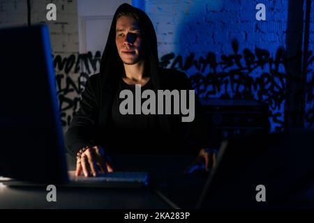 Portrait of hidden hacker man wearing sweatshirt with hood engaged in hacking into security systems, sitting in dark basement room with blue neon Stock Photo